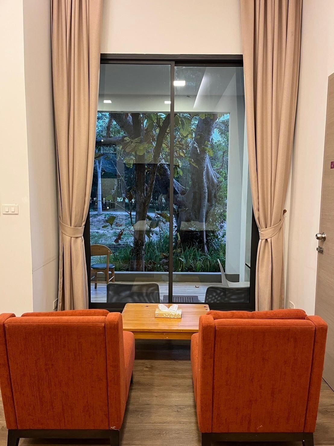 A room with a large window, two orange chairs, a wooden table, and a view of trees and a garden outside.