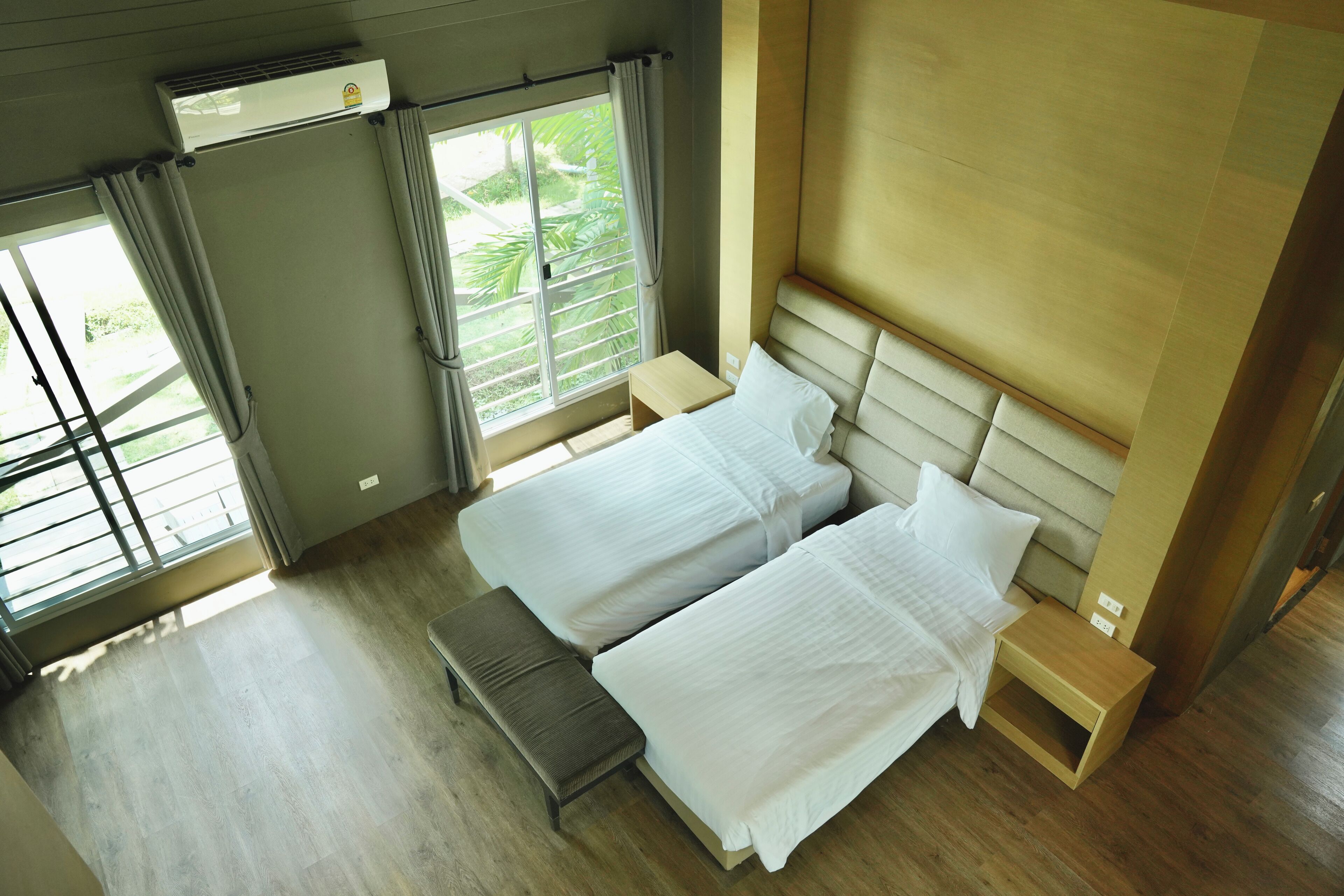 A hotel room with two beds, a bench, a small table, an air conditioning unit, and a window with curtains.