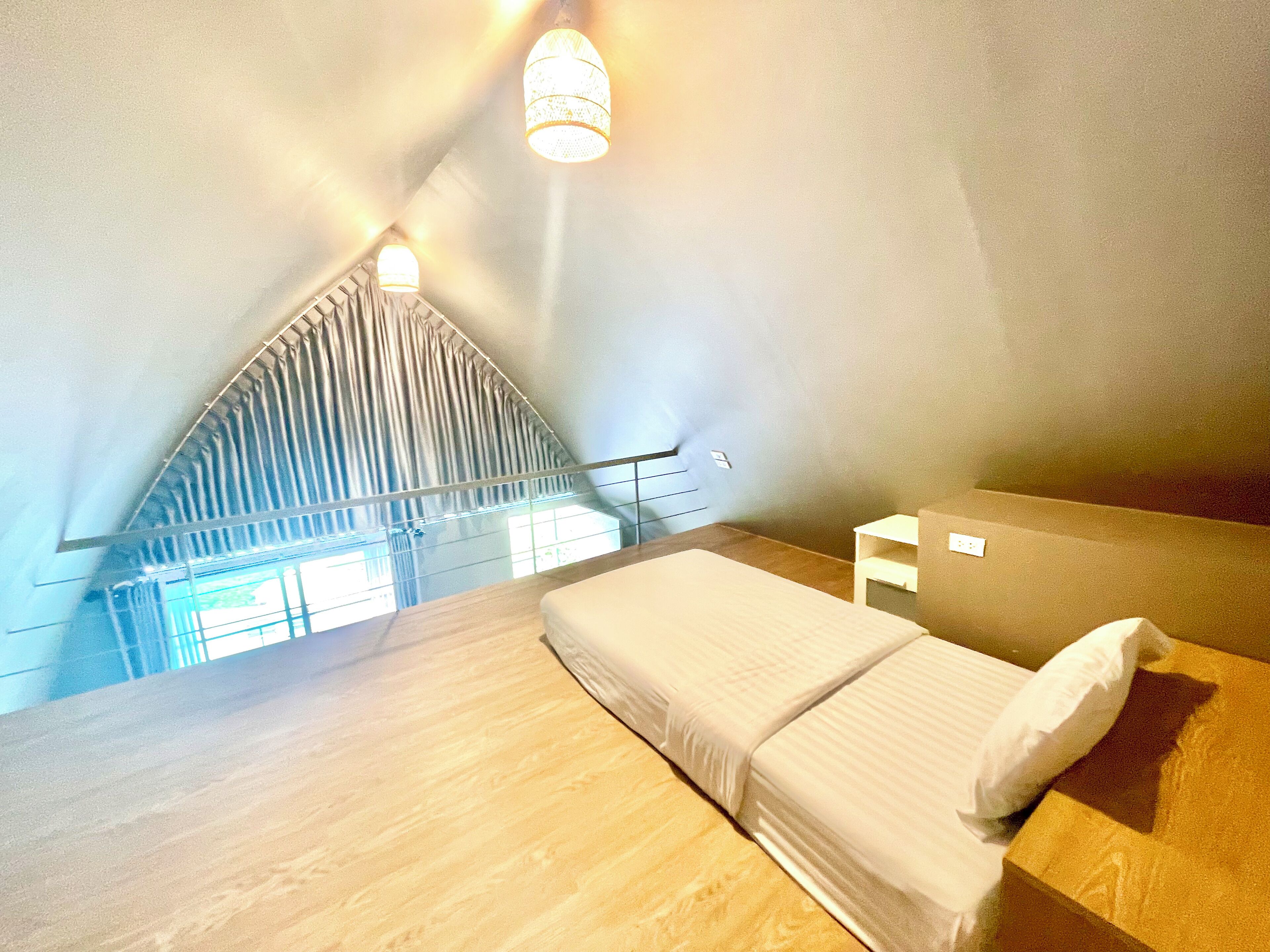 A high-ceilinged room with a sloped ceiling, a bed with a white mattress and pillow, a small bedside table, and a window with curtains.