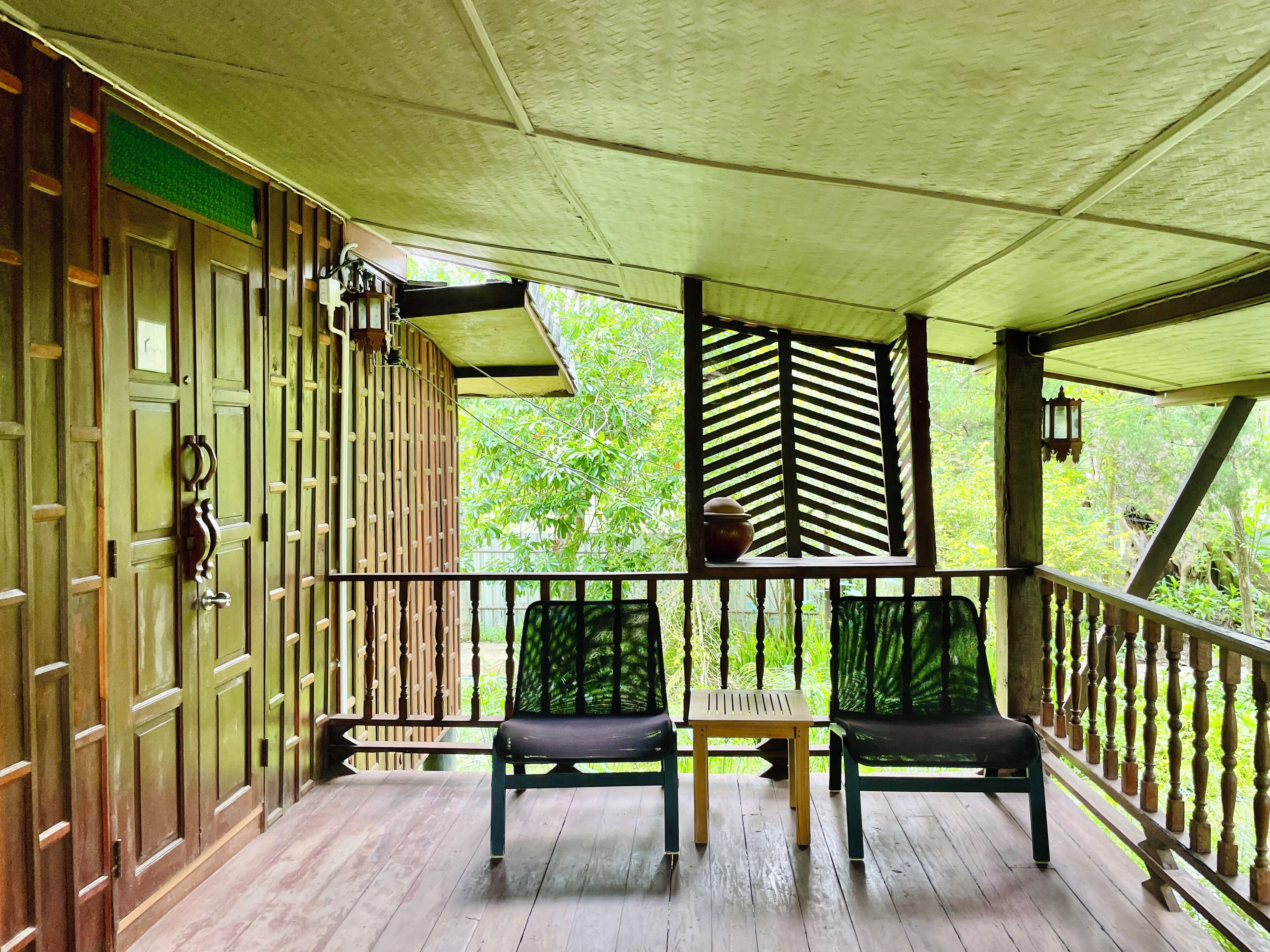 A wooden porch with two chairs and a table, a green door, and a view of greenery through a window.