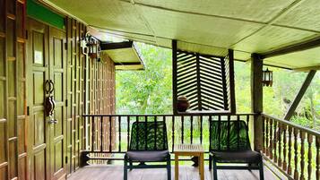 A wooden porch with two chairs and a table, a green door, and a view of greenery through a window.