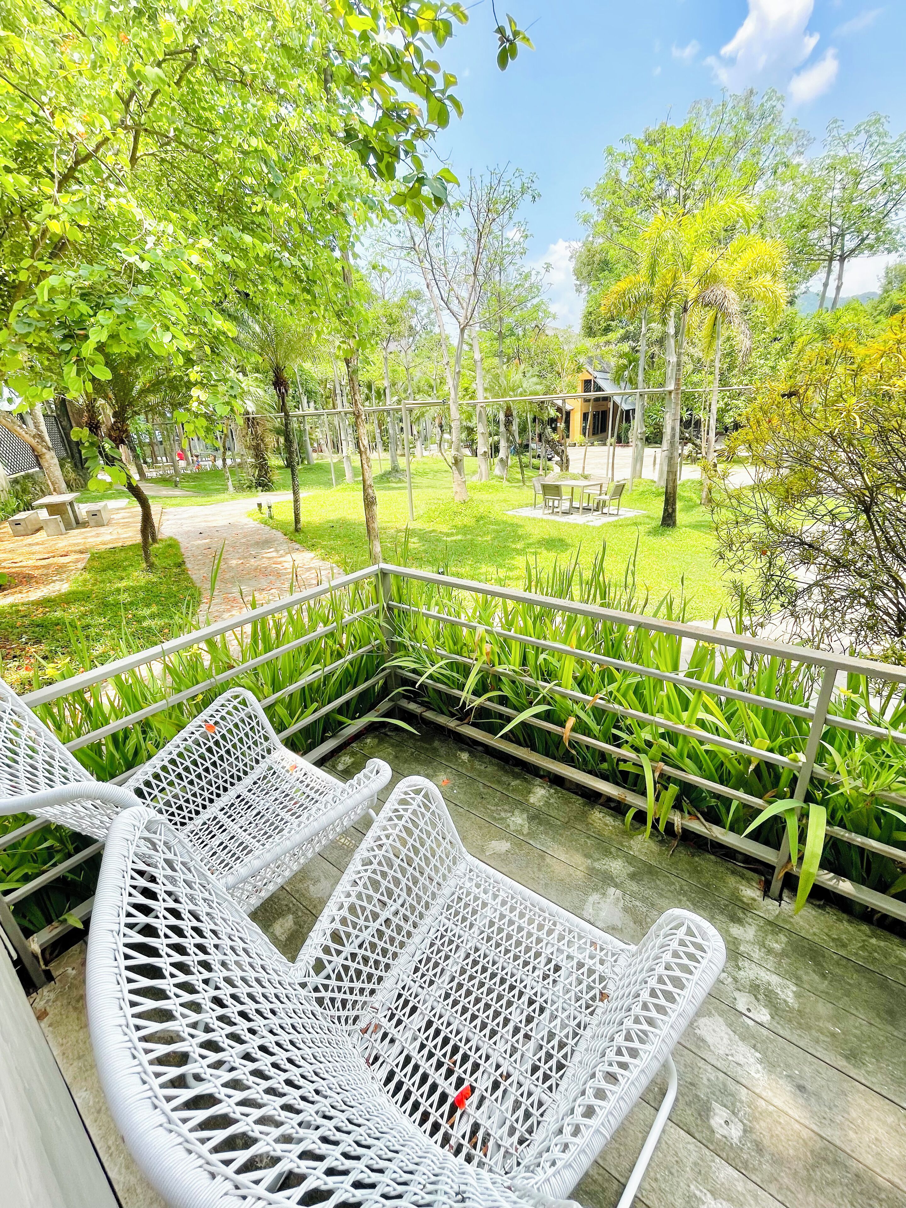 A covered outdoor seating area with white wicker chairs, surrounded by lush greenery and a clear sky.
