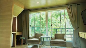A room with two armchairs, a small table, and a stool. There is a large window with curtains, and a TV mounted on the wall.