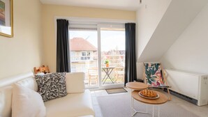 A modern living room with a white sofa, a small wooden table with a plate of cookies, a painting on the wall, and a view of the outside balcony. - Lovely apartment for 4 guests with pool, WIFI, TV and balcony (De Haan)