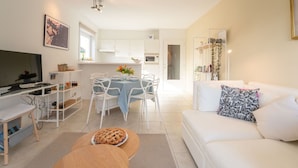 A modern living room with a white sofa, a coffee table, and a TV on a stand. There is a dining area with a table and chairs, and a kitchen in the background. - Lovely apartment for 4 guests with pool, WIFI, TV and balcony (De Haan)