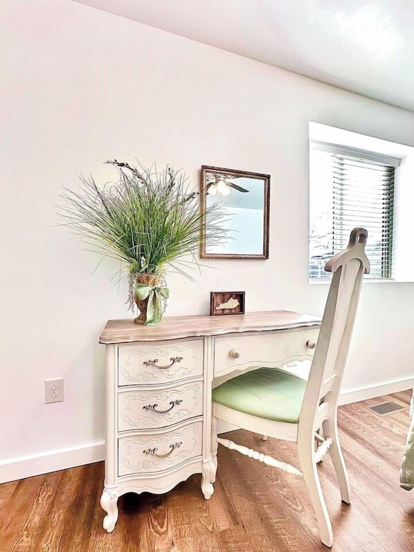 A room with a wooden desk, a chair, a vase with green plants, a framed picture, and a window with blinds.