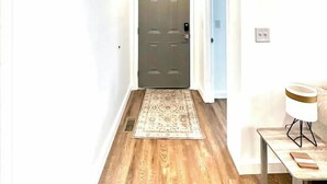 Un pasillo con piso de madera, alfombra estampada, una puerta con cerradura electrónica y una pequeña mesa con lámpara.