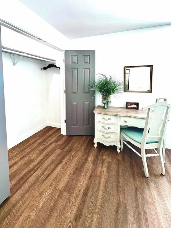 Una habitación con suelo de madera, un escritorio con cajones, una silla, una puerta y una planta.