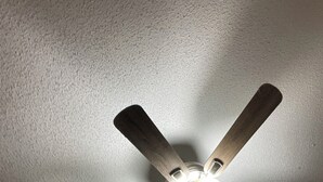 Un ventilador de techo con cuatro aspas y una lámpara en una habitación con paredes blancas y una puerta visible al fondo.