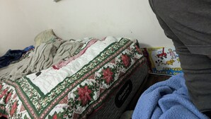 Una cama con colcha floral, una manta gris y una toalla azul. Se ve una ventana con persianas y un libro titulado “Art Pad” sobre la cama.