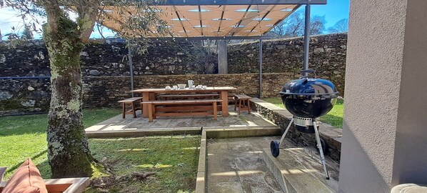 An outdoor patio with a wooden table and chairs, a barbecue grill, and a stone wall in the background.