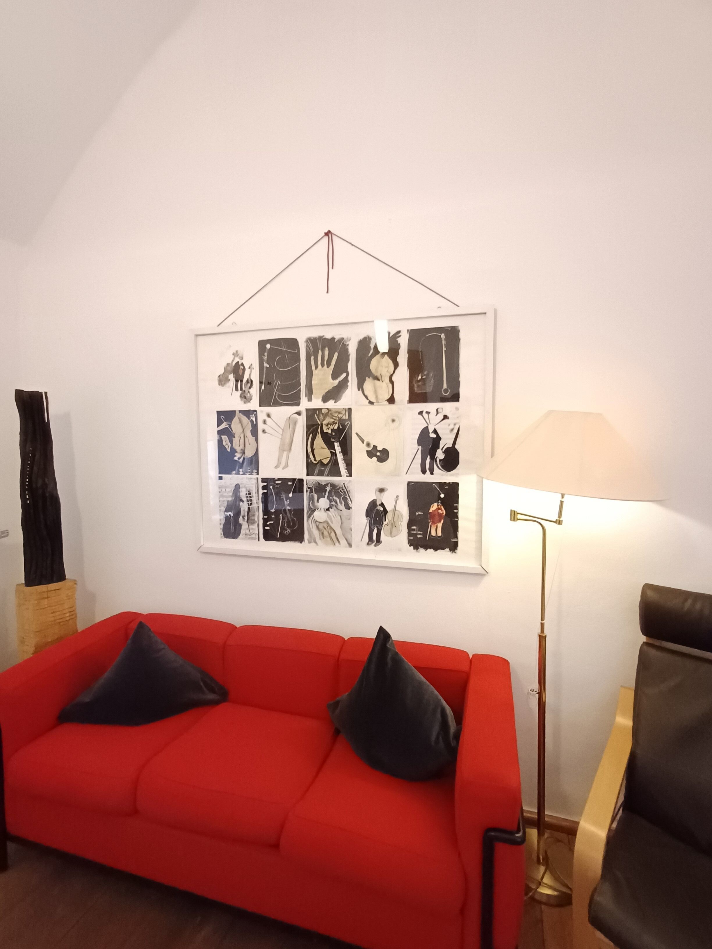En röd soffa med svarta kuddar, en inramad collagebestående av svartvita bilder på väggen och en golvlampa.