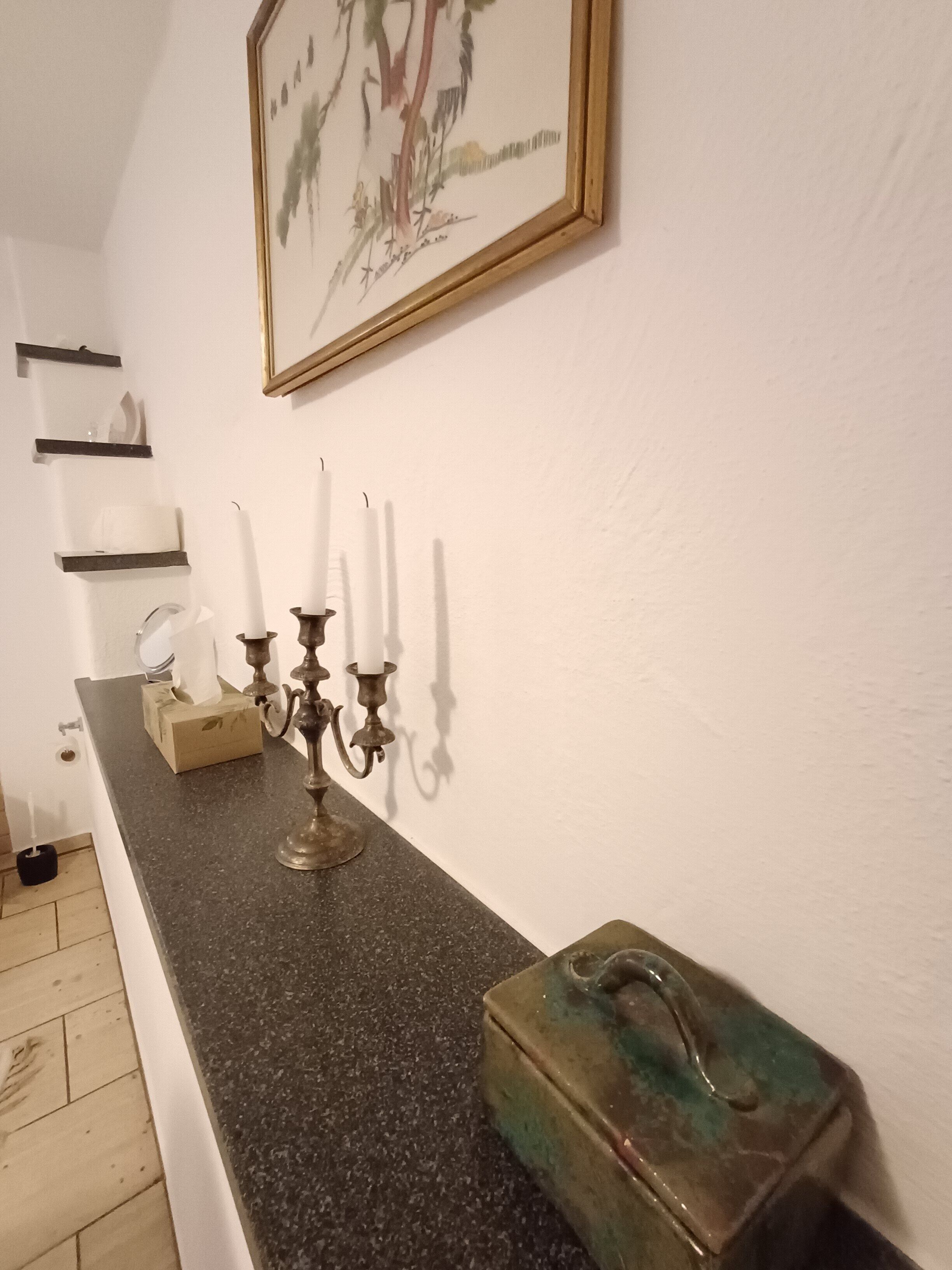 Un pasillo con encimera de granito oscuro, un candelabro de bronce con velas blancas, un cuadro enmarcado y una caja metálica verdosa.