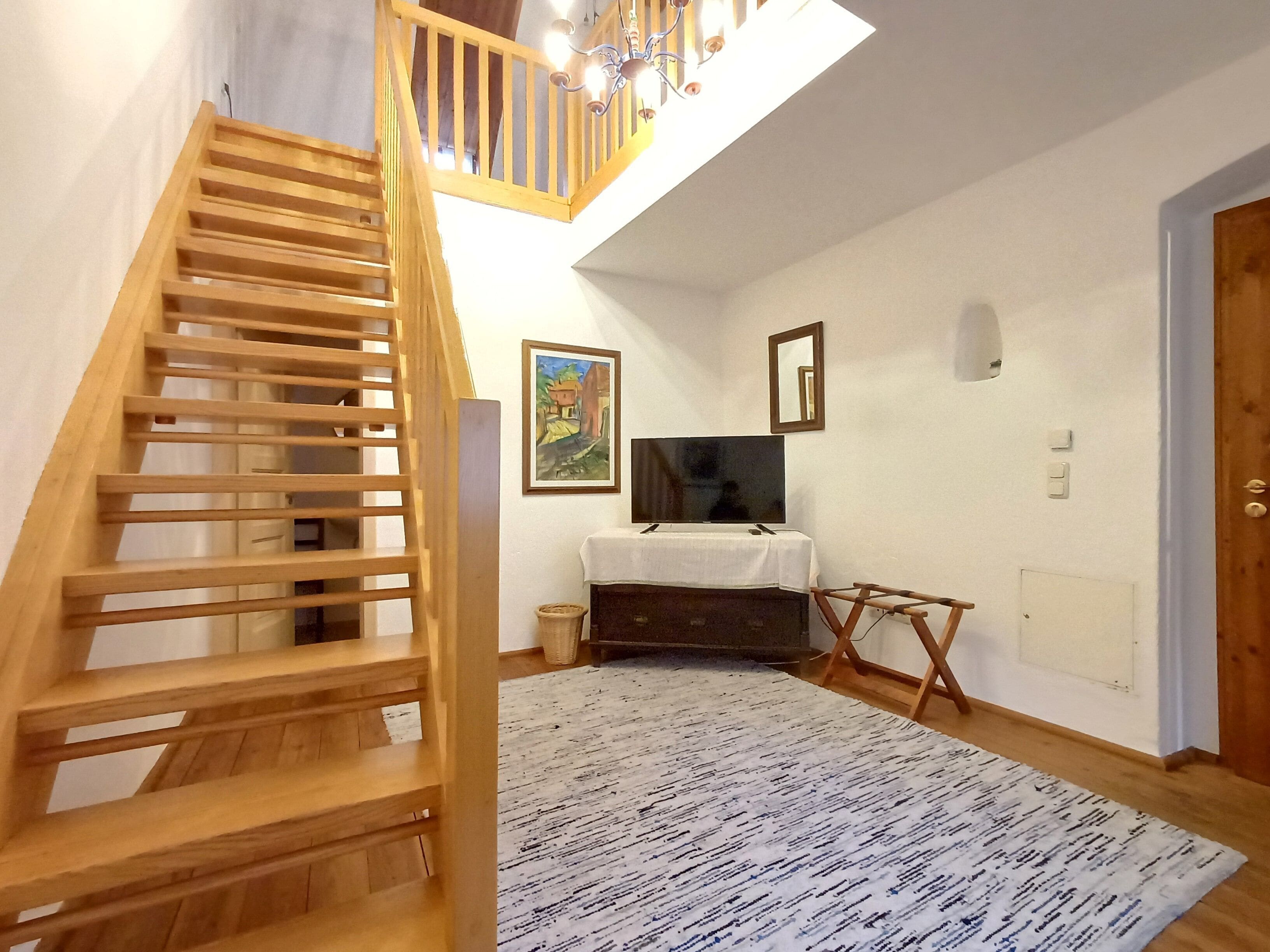 Una escalera de madera conduce a un entrepiso con techo blanco y un candelabro. Un televisor de pantalla plana está montado en la pared, y se observa una pequeña mesa con mantel blanco.