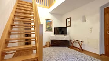 Una escalera de madera conduce a un entrepiso con techo blanco y un candelabro. Un televisor de pantalla plana está montado en la pared, y se observa una pequeña mesa con mantel blanco.