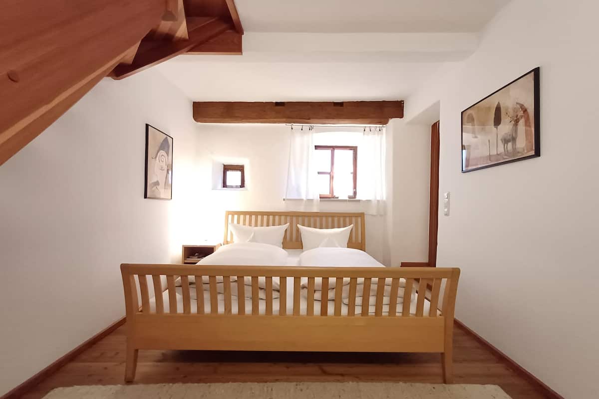 Ein Schlafzimmer mit einem hölzernen Bett, einem kleinen Fenster mit Vorhängen, einem gerahmten Bild an der Wand und einem Holzbalken an der Decke.