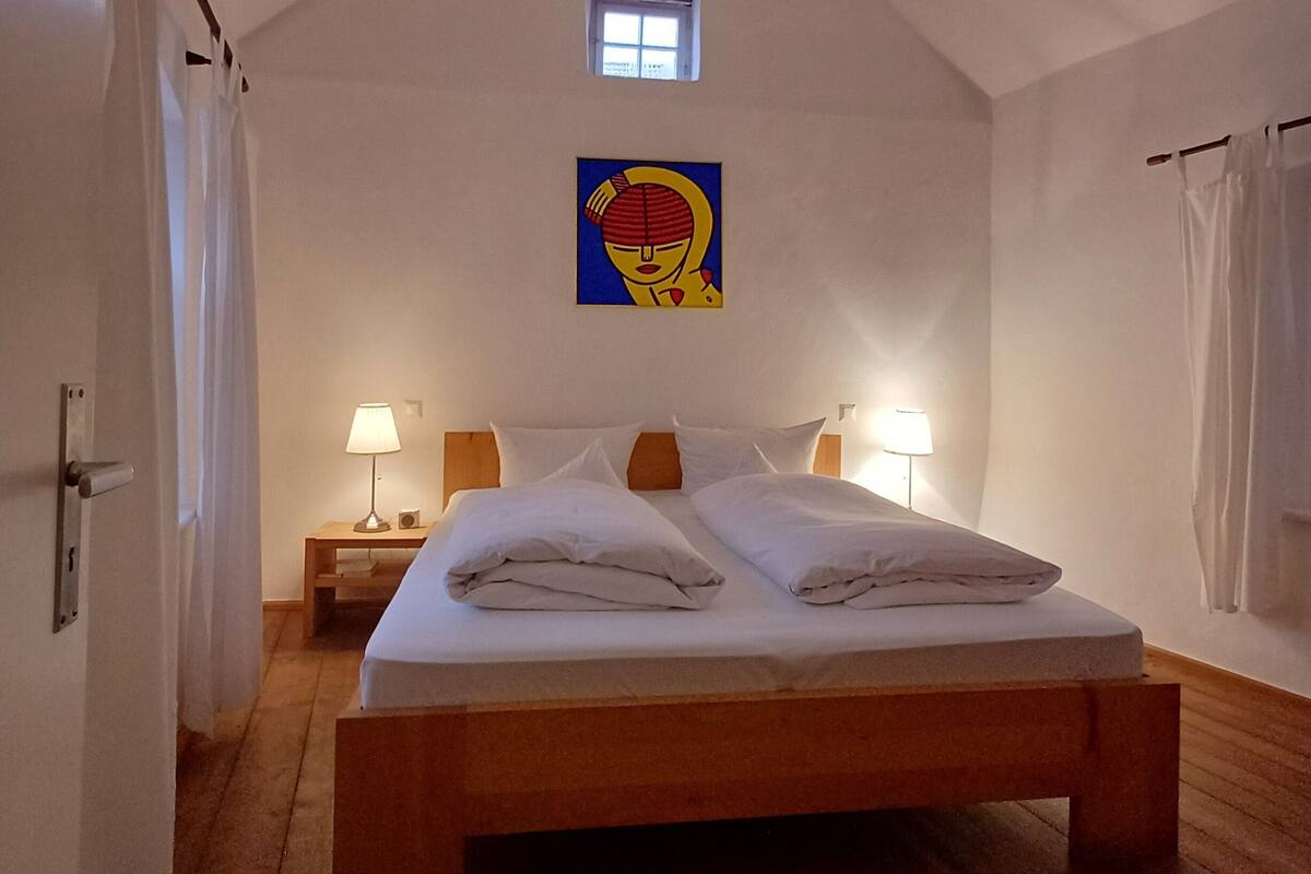 Ein Schlafzimmer mit schrägem Dach, einem Bett mit weißer Bettwäsche, zwei Nachttischlampen, einem kleinen Fenster und einem Bild an der Wand.