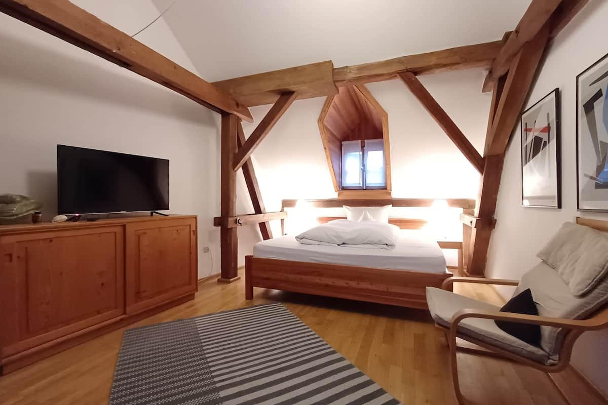 Ein Schlafzimmer mit einem hölzernen Bett, einem Fernseher auf einer Holzkommode, einem Fenster mit Vorhängen und einem Stuhl.