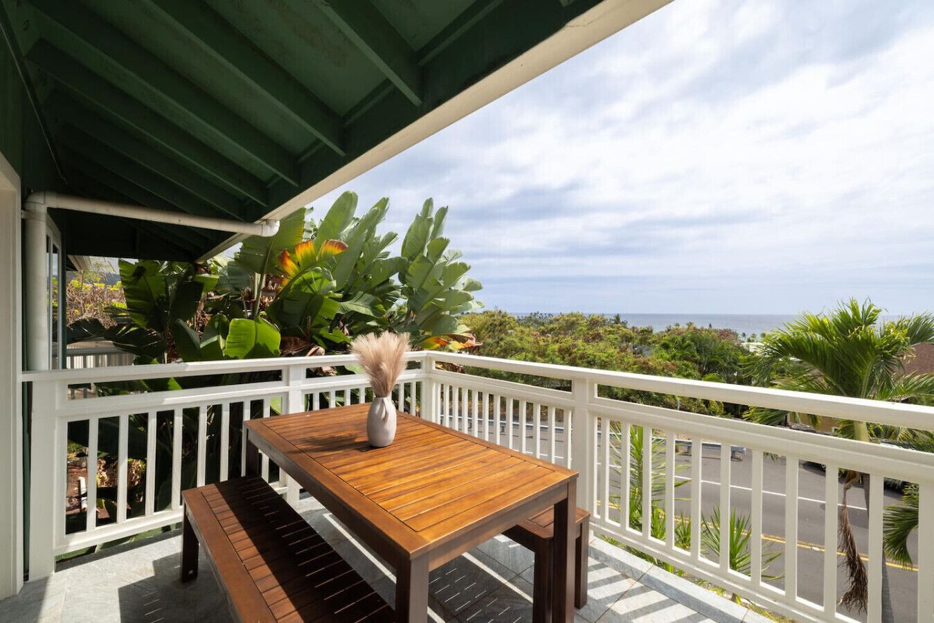 Ein Holztisch mit einer Vase auf einem Balkon mit weißen Geländern, mit Blick auf eine Küstenlandschaft mit Vegetation und Meer.