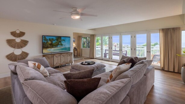 A spacious living room with a large gray sofa, a flat-screen TV, and a ceiling fan. - Kona Sunset Retreat | Walk to Shops & Dining (Kailua-Kona)