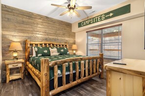 Una cama con estructura de madera, un edredón verde y dos almohadas, una mesita de noche con una lámpara, una ventana con persianas, y un cartel que indica 'Campsites'.