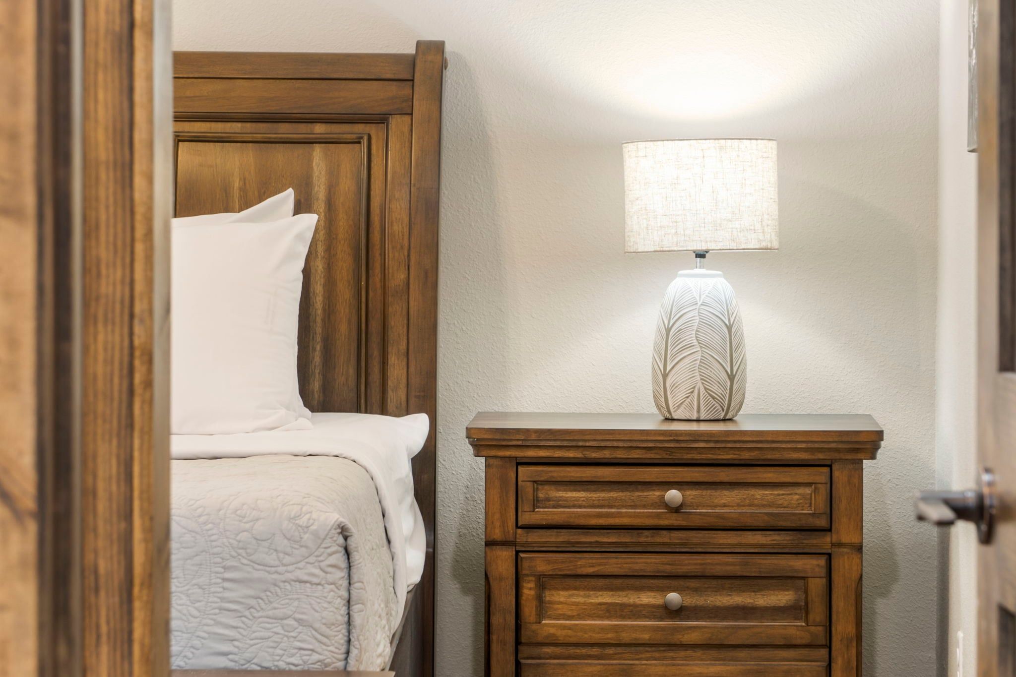 A bedroom with a wooden headboard, a bedside table with a lamp, and a bed with white bedding.
