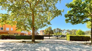 Un parking clos, avec un arbre au premier plan, une clôture verte et des bâtiments en arrière-plan.