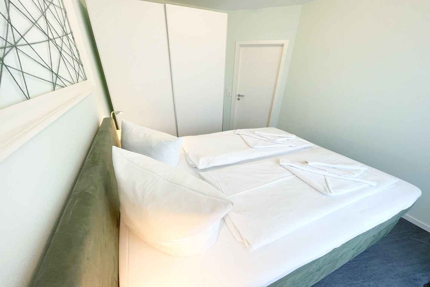 A neatly made bed with white linens and pillows, a wooden headboard, and a door in the background.