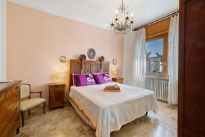 Une chambre à coucher comprenant un lit, une commode en bois, une chaise, une table de chevet, un lustre et une fenêtre avec des rideaux.