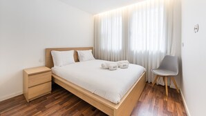 A modern bedroom with a wooden bed, a bedside table, a chair, and sheer curtains. - GuestReady - Ceuta Axis Awaits (Porto)