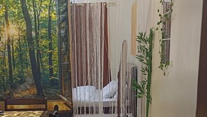 A room with a dining table and chairs, a bed with a white bedspread, a curtain with vertical lines, a wall-mounted plant, and a television.