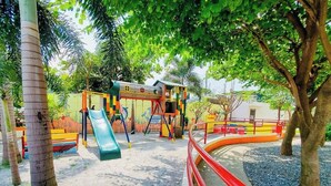 A playground with a slide and climbing structures, surrounded by trees and a red safety barrier.