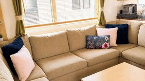 Un salon avec un canapé d’angle beige, une table basse en bois et une fenêtre équipée de stores.