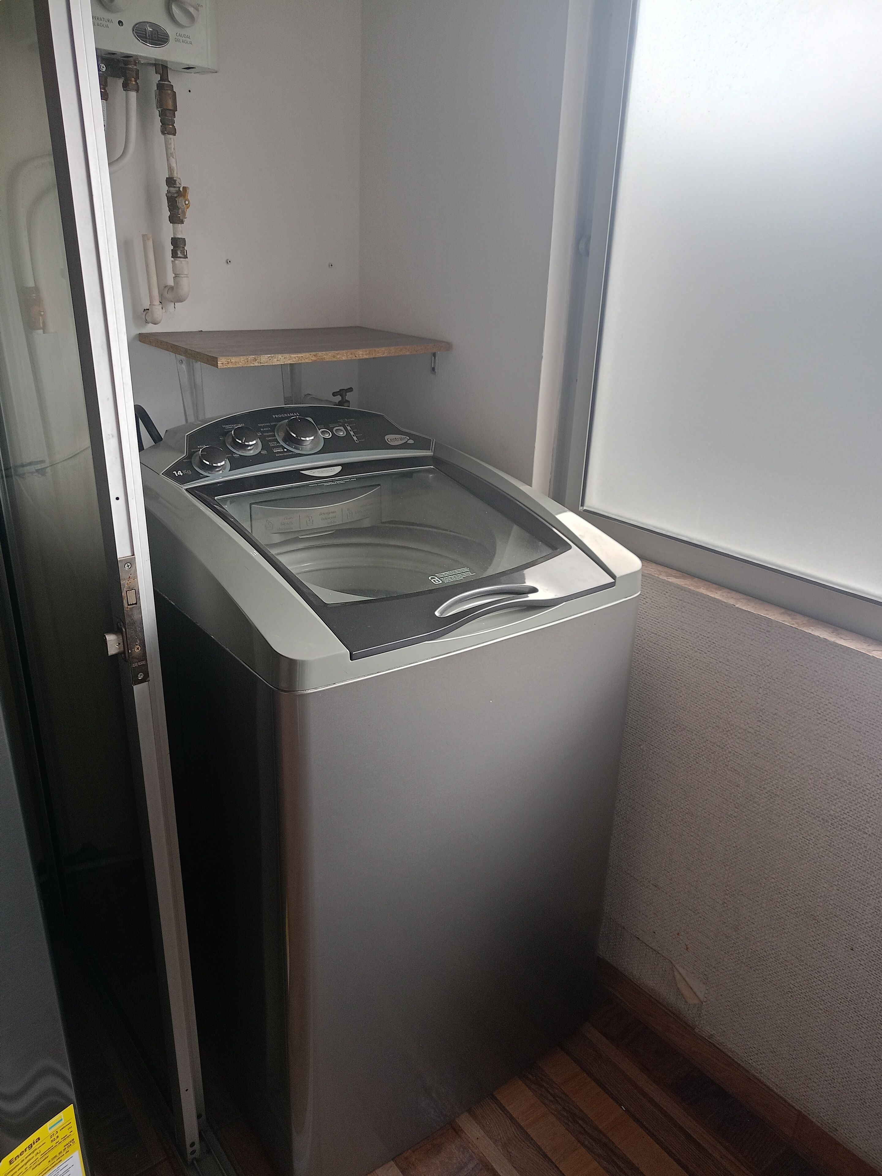 A compact, stainless steel front-loading washer and dryer unit in a small laundry room with a wooden shelf above it and a window to the right.