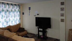 A living room with a sofa, a flat-screen TV mounted on the wall, and a window with curtains.