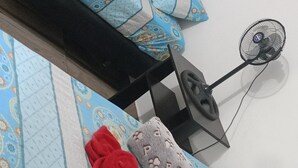 A bedroom with a patterned bedspread, pillows, a bedside table, and a wall-mounted fan.