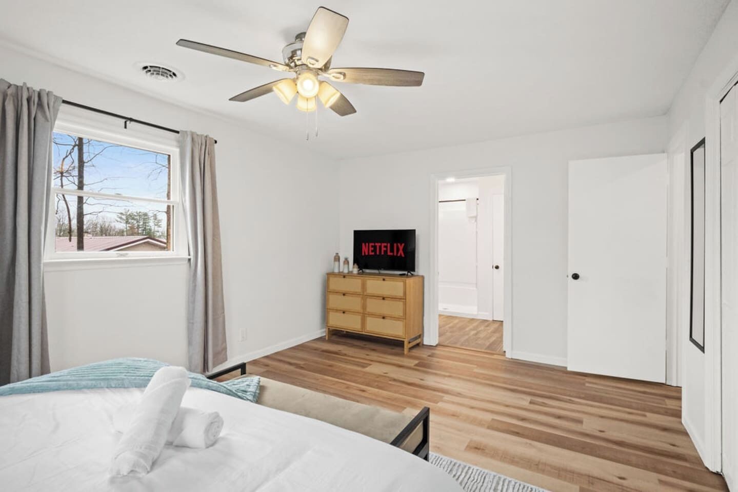 A bedroom with a bed, a TV on a wooden dresser, a ceiling fan, and a window with curtains.