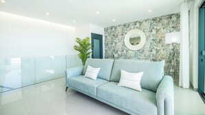 A modern living room with a light blue sofa, white cushions, a round mirror, and a floral-patterned wall. - Sob O Mar II - Stunning beach/sea view (Armação de Pêra)