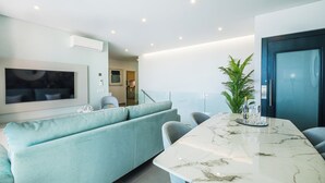 A modern living room with a light blue sofa, a marble dining table, and a large flat-screen TV mounted on the wall. - Sob O Mar II - Stunning beach/sea view (Armação de Pêra)