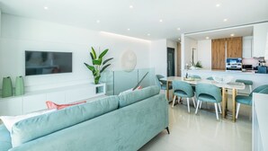 A modern living room with a sofa, a dining table with chairs, a TV mounted on the wall, and a kitchen area visible through an open door. - Sob O Mar III - Stunning beach/sea view (Armação de Pêra)