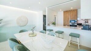 A modern kitchen with wooden cabinets, a central island with stools, and a dining area with a marble table and green chairs. - Sob O Mar III - Stunning beach/sea view (Armação de Pêra)