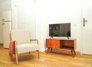 A room with a wooden floor, a beige armchair with a red throw, a wooden TV stand with a flat-screen TV, and a white door with a brass handle.