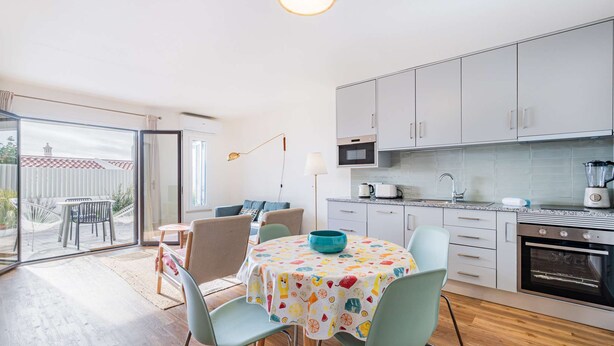 A modern kitchen with white cabinets, a microwave, and a stove. There is a dining area with a table and chairs, and a large window providing a view of the outside. - Stanchion Apartment (Albufeira)