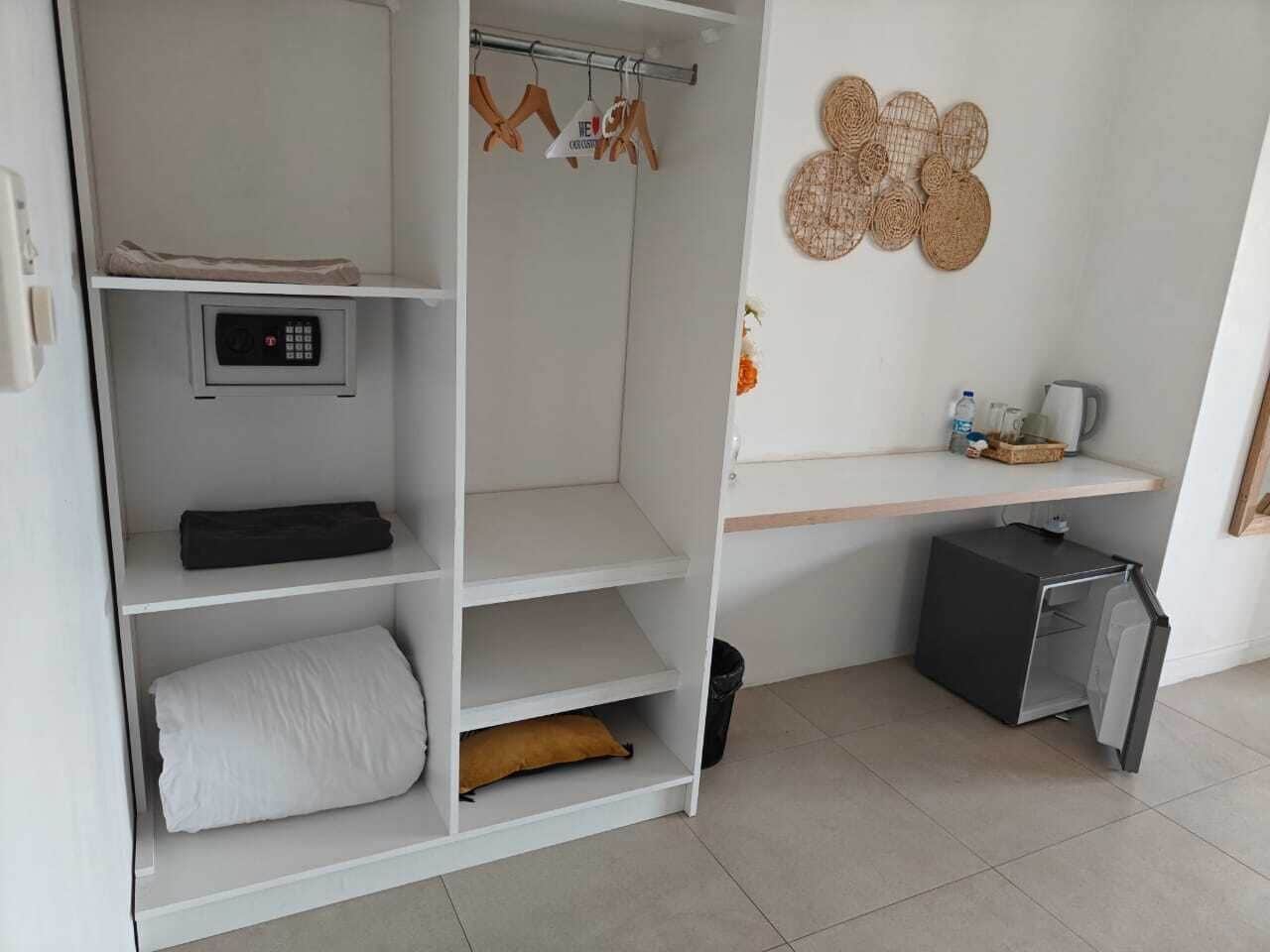 A bedroom with a wardrobe, a small shelf with toiletries, a decorative wall hanging, and a compact refrigerator.