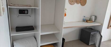 A bedroom with a wardrobe, a small shelf with toiletries, a decorative wall hanging, and a compact refrigerator.