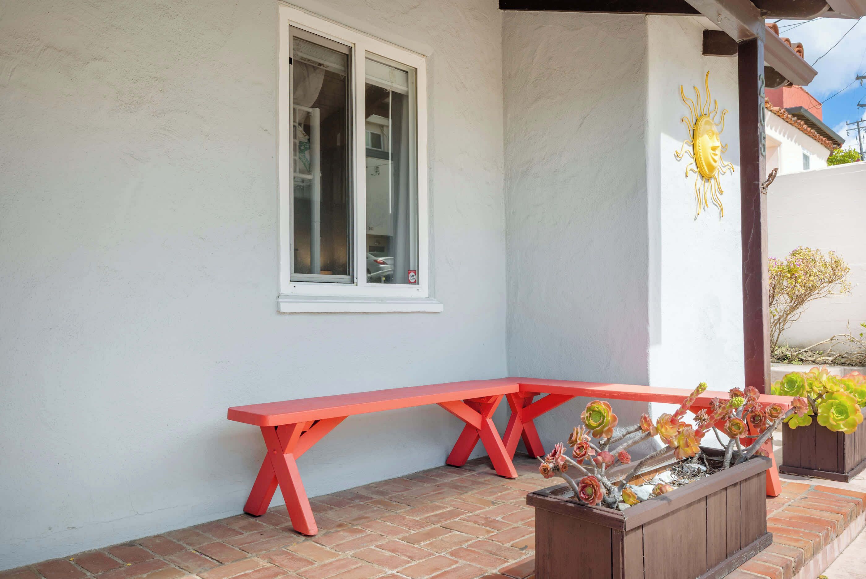 A red bench outside a building with a window and a planter with flowers.