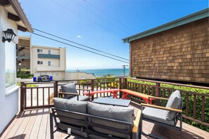 A wooden deck with outdoor furniture, a view of the sea, and a building with a balcony. - Adorable Rio Del Mar Beach House - Steps to beach! (Aptos)