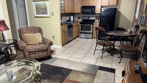 A compact kitchen with wooden cabinets, a microwave, an oven, and a refrigerator. There is a dining area with a table and chairs, a patterned armchair, and a glass coffee table.