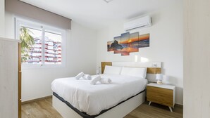 Habitación de hotel moderna con una cama grande, una mesita de noche con lámpara, aire acondicionado de pared y un amplio ventanal con vistas a edificios.
