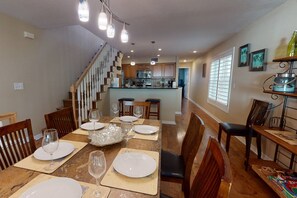 Comedor con una gran mesa dispuesta para seis personas, una cocina al fondo y una escalera que sube a otro nivel.
